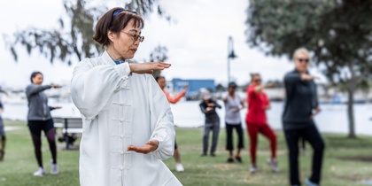 En grupp människor utövar tai chi utomhus i en park, med en kvinna i vit dräkt i förgrunden.