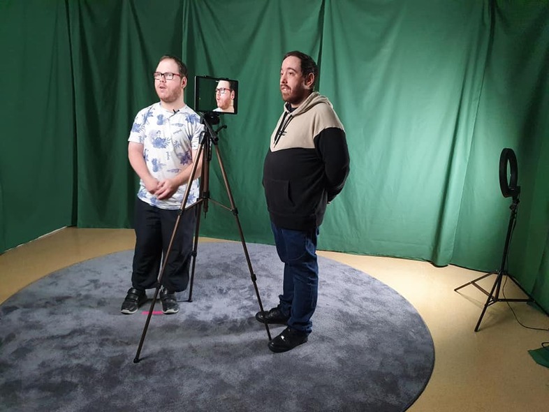 Två män står i en studio med gröna draperier, en kamera på stativ och ringlampa syns.