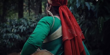 En person i grön traditionell klädsel och röd sjal bär en trumma i en skogsmiljö. Text: Budakattu, Archive of Tribal Musical Arts.