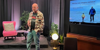 Man står på en scen med svart bakgrund, bredvid en TV-skärm med text och en bild på två personer vid havet.