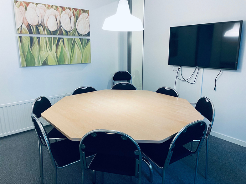 Ett mötesrum med åttakantigt bord, åtta stolar, vägg-TV, tavla med blommor och vit lampa.
