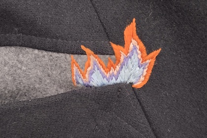 Närbild på en svart tygficka med en broderad eldflamma i orange och blått vid öppningen.