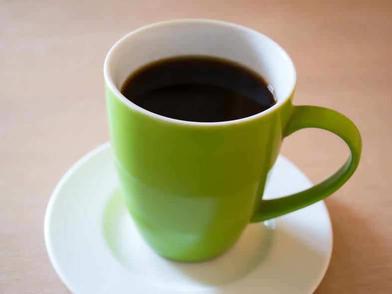 En grön kopp med svart kaffe står på ett vitt fat på ett ljust bord.