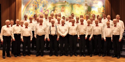 En stor manskör i vita skjortor och röda flugor står uppställda framför en färgglad målning.