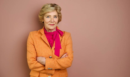 Kvinna med blont hår i orange kavaj och cerise scarf står med armarna i kors mot en rosa vägg.