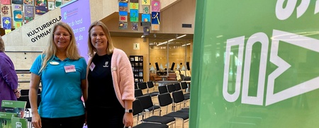 Två kvinnor står vid ett informationsbord på en mässa i en ljus lokal med färgglada tavlor på väggen.