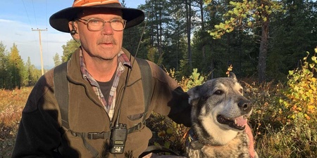 En person i friluftskläder sitter på knä i skogen bredvid en glad hund under en solig dag.