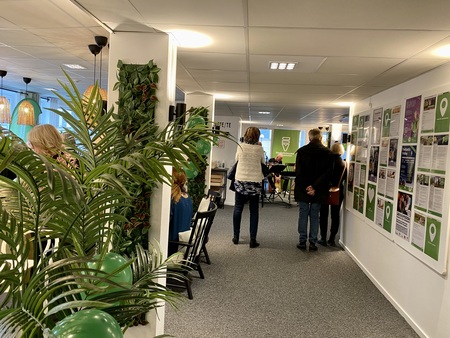 Inomhusmiljö med människor som står och sitter, gröna dekorationer och informationsaffischer på väggen.