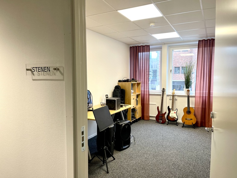 Ett ljust musikrum med skrivbord, dator, högtalare och tre gitarrer vid fönstret.