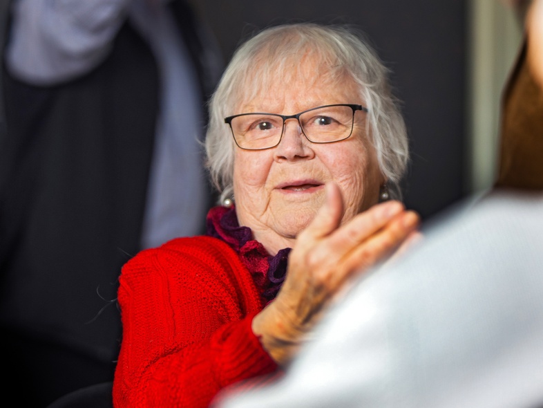 Äldre kvinna med glasögon och röd tröja sitter inomhus och gestikulerar med handen, andra personer i bakgrunden.