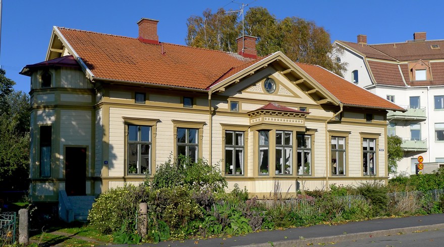 Gult trähus med rött tegeltak och stora fönster, omgiven av grönska och en asfalterad väg framför.