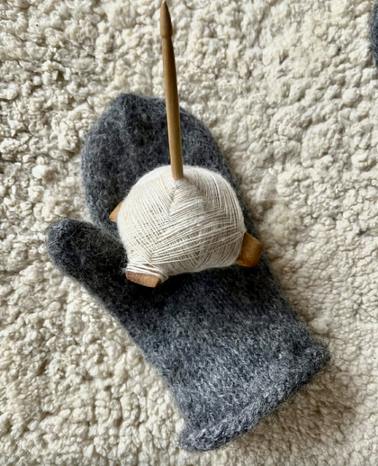 En grå stickad vante ligger på en ljus, fluffig matta med en slända och garnnystan ovanpå.