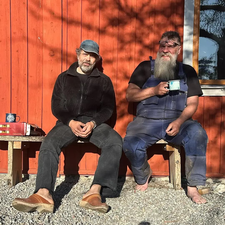 Två män sitter på bänkar utomhus mot en röd trävägg, en dricker kaffe, båda ser avslappnade ut.