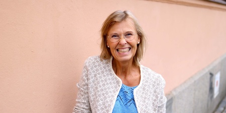 Leende äldre kvinna med glasögon står mot en ljusrosa vägg utomhus.