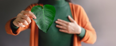 Kvinna håller ett grönt hjärtformat blad framför bröstet, klädd i grön tröja och orange kofta.