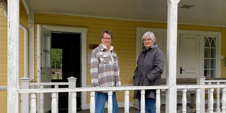 Två äldre kvinnor står på en veranda framför ett gult trähus med vita detaljer.