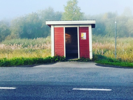 En liten röd busskur står vid en landsväg med dimmig natur i bakgrunden.
