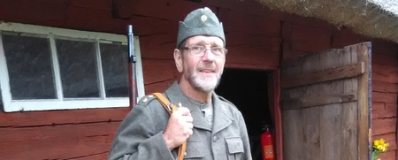 Man i militäruniform står utanför en röd träbyggnad med ett gevär på axeln.