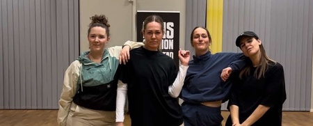 Fyra personer poserar tillsammans i en dansstudio med trägolv och gråa gardiner.