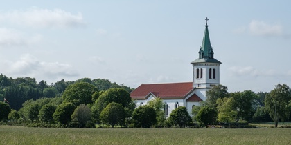 En vit kyrka med rött tak och grön spira står omgiven av träd på en äng under blå himmel.