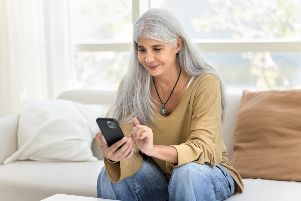 Kvinna med långt grått hår sitter i soffa och använder en smartphone.