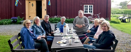 En grupp vuxna sitter runt ett bord utomhus framför ett rött trähus och fikar tillsammans.