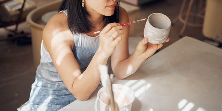 En kvinna målar en keramikmugg med pensel i en ljus keramikstudio.