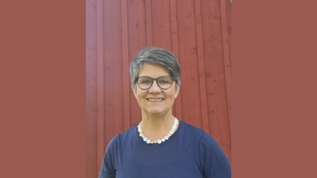 Kvinna med kort grått hår, glasögon och pärlhalsband står framför en röd trävägg och ler.