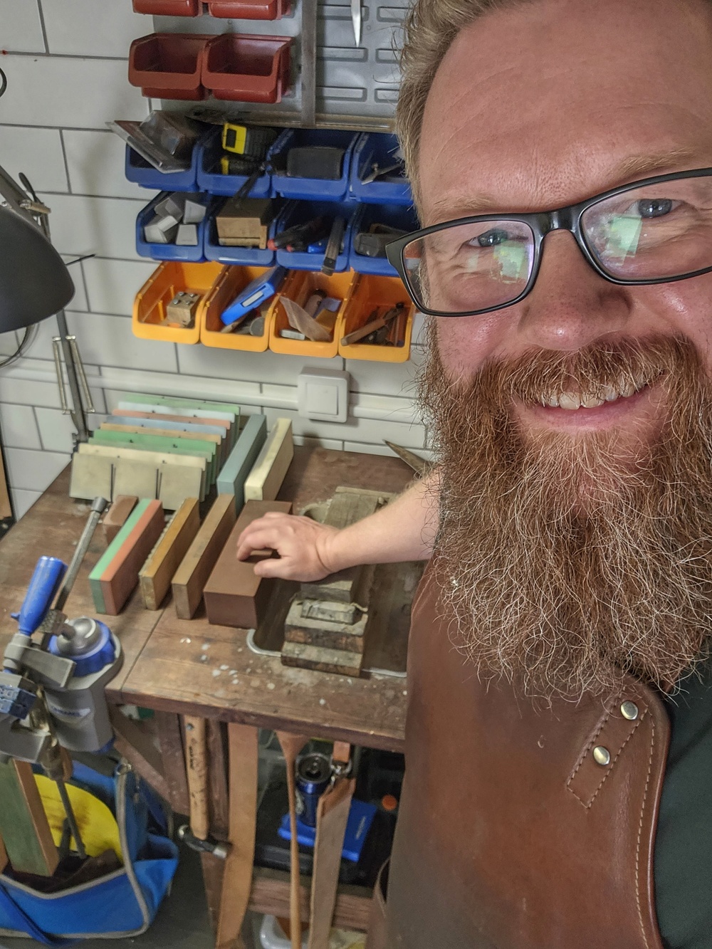 Man med skägg och glasögon står vid arbetsbänk med slipstenar och verktyg i verkstad.