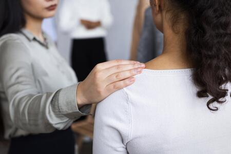 En person lägger en hand tröstande på axeln av en annan person i en inomhusmiljö.
