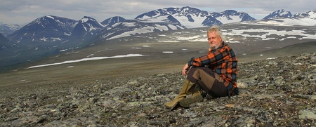 En man i rutig skjorta sitter på en stenig fjällsluttning med snötäckta berg i bakgrunden.