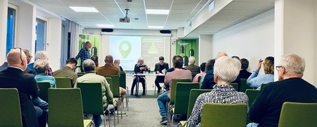 Ett möte eller seminarium med vuxna deltagare sittande i gröna stolar i ett konferensrum.