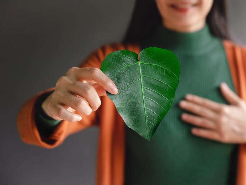 Kvinna håller ett grönt hjärtformat blad framför bröstet, klädd i grön tröja och orange kofta.