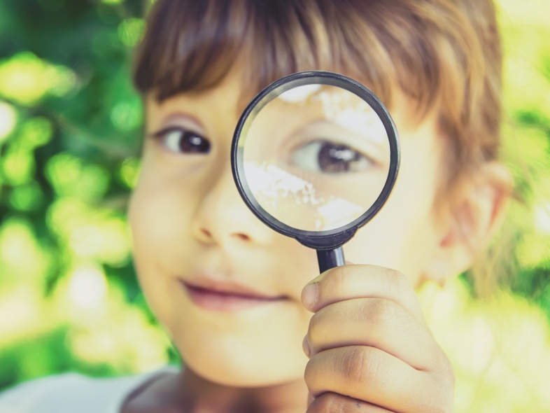 Ett barn håller upp ett förstoringsglas framför sitt öga utomhus, med grönskande bakgrund.