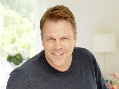 Man med kort ljust hår och mörk tröja sitter avslappnat inomhus och ler mot kameran.