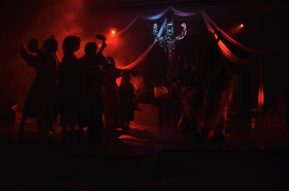 Människor dansar på en scen med röd belysning och dekorationer i bakgrunden.