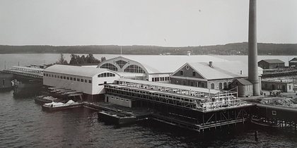 Svartvit bild av en industribyggnad vid vattnet med stor skorsten och bryggor.