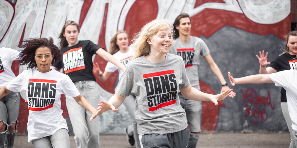 En grupp unga personer dansar energiskt utomhus framför en graffitivägg, alla bär t-shirts med texten 'DANS STUDIO'.