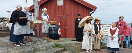 En grupp människor i historiska kläder står utomhus vid en röd träbyggnad, några samtalar och andra tittar på.
