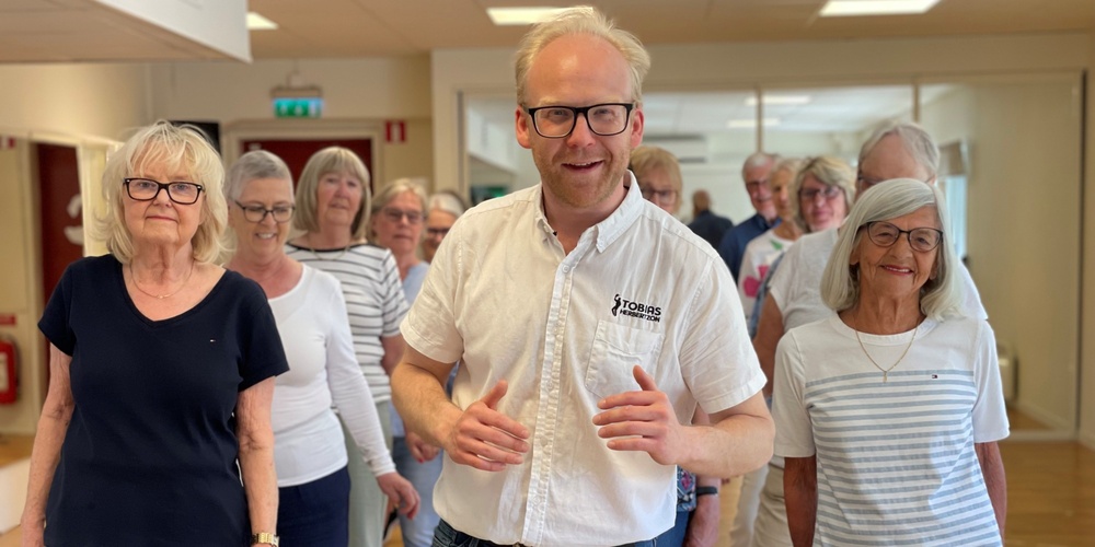 En grupp äldre personer och en yngre man står tillsammans inomhus, leende och avslappnade.