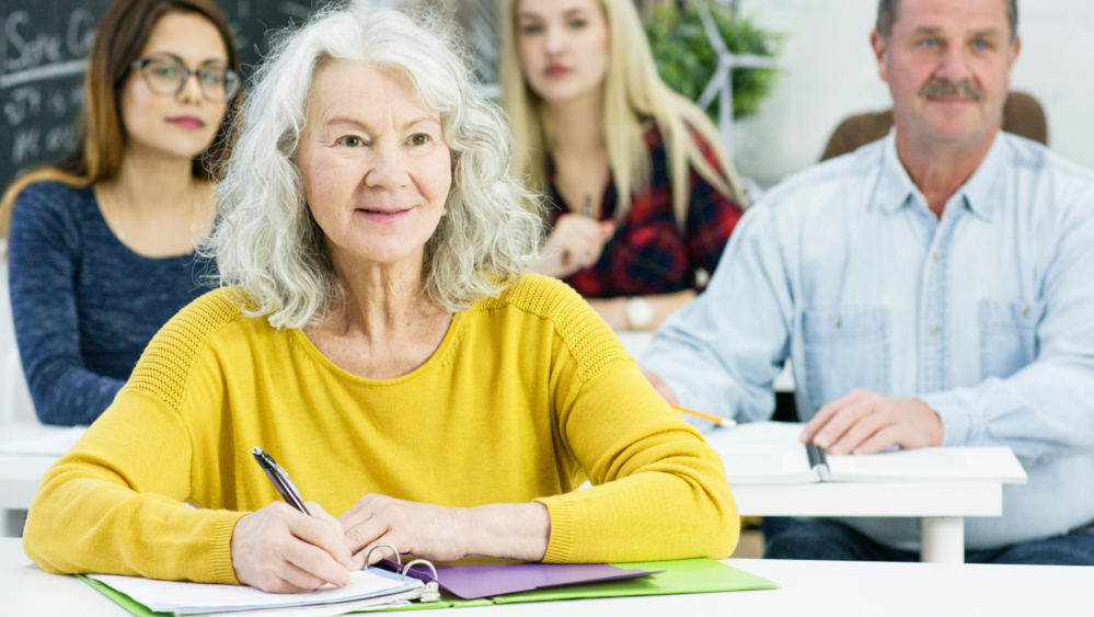 Äldre kvinna i gul tröja, sitter vid bänk och lyssnar på föreläsning