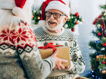 Två äldre personer i jultröjor och tomteluvor utbyter en inslagen present framför en julgran.