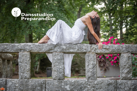 Kvinna i vit klänning poserar dansande på en stenmur utomhus med grönska och blommor i bakgrunden.