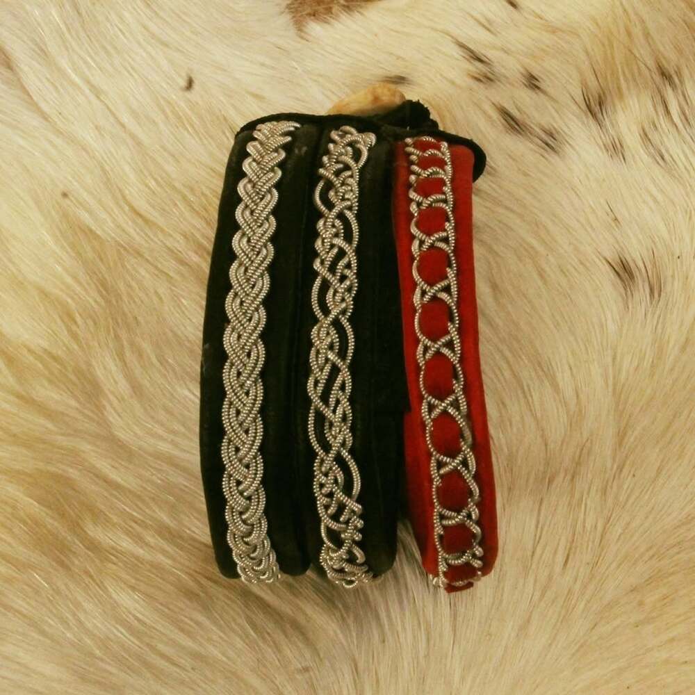 Tre armband i svart och rött med silverfärgade mönster ligger på ljust djurpälsunderlag.