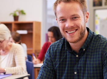 Leende man sitter vid skolbänk i klassrum, skriver i anteckningsbok. Andra vuxna studenter i bakgrunden.