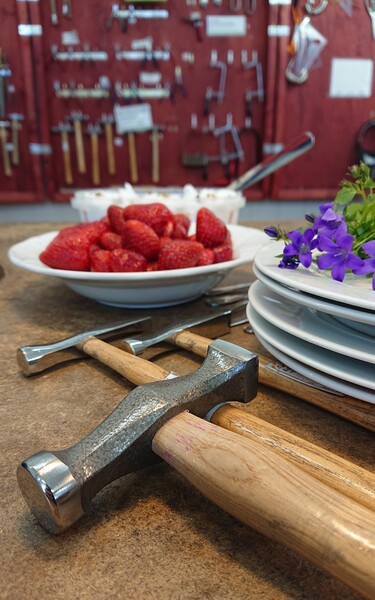 En närbild av hammare, tallrikar, jordgubbar och blommor på ett bord i en verkstadsmiljö.