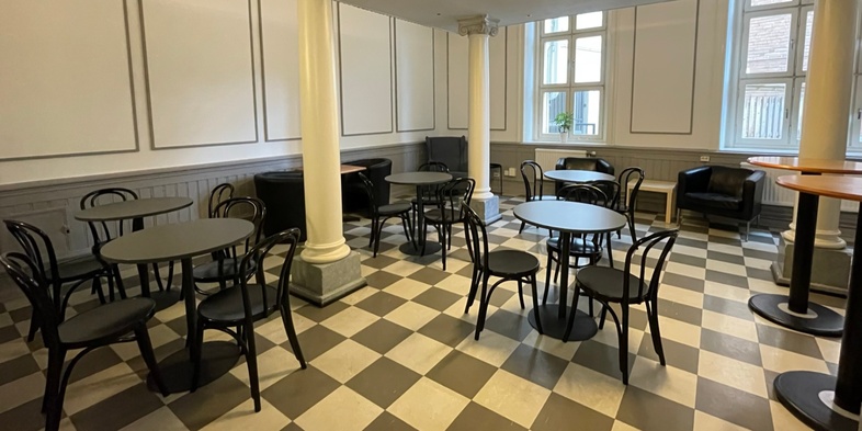 Ett tomt café eller uppehållsrum med svartvita rutiga golv, runda bord, svarta stolar och stora fönster.