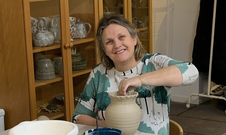 Kvinna ler och arbetar med keramik vid ett bord, omgiven av hyllor med keramikföremål.