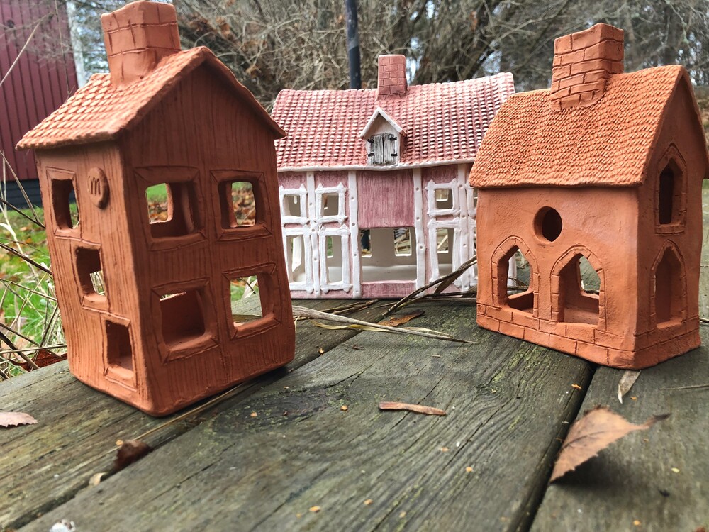 Tre små modellhus i lera och trä står på en väderbiten träyta utomhus med gräs och träd i bakgrunden.