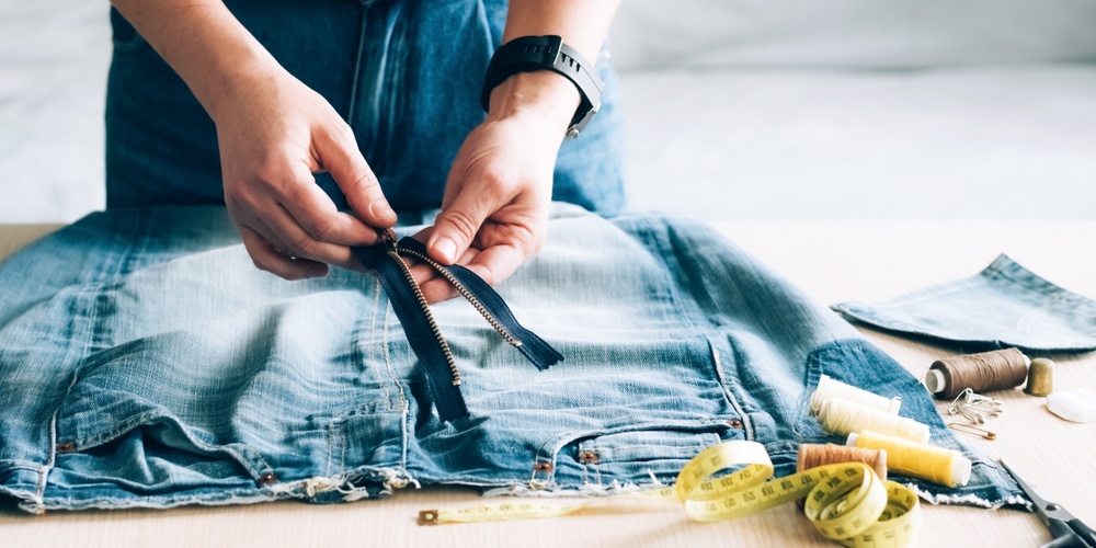 Person arbetar med att sy om ett par jeans, med dragkedja, måttband och sytråd på bordet.